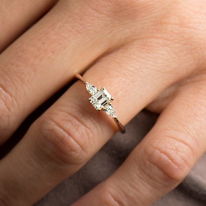 May include: A delicate gold ring with a square-shaped center stone and smaller stones on either side. The ring is on a person's finger.