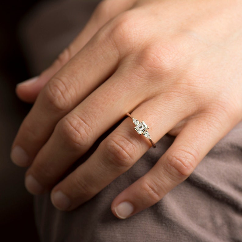 May include: A delicate gold ring with a square-shaped center stone and smaller stones on either side. The ring is on a person's finger.