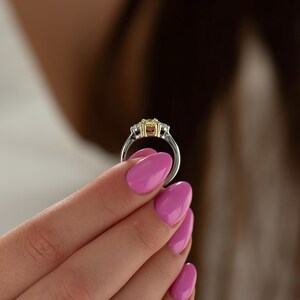 K&ouml;nnte beinhalten: Ein silberner Ring mit einem gelben und wei&szlig;en Edelstein in der Mitte. Der Ring wird von einer Hand mit rosafarbenem Nagellack gehalten.
