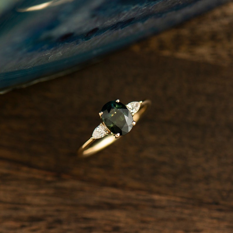 May include: A gold ring with a dark green oval gemstone and two smaller pear-shaped diamonds on either side.