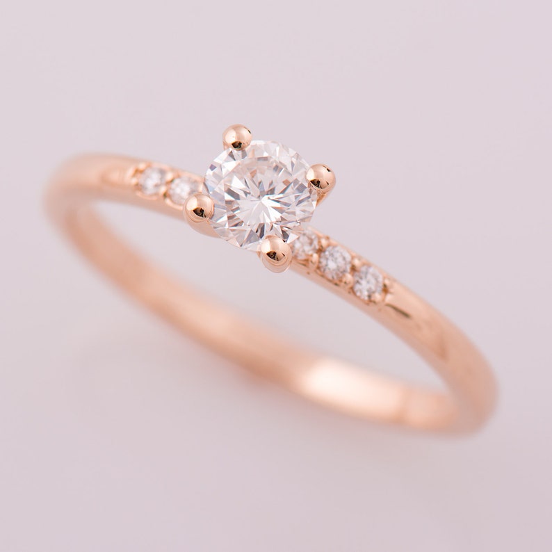 Peut inclure: Bague en or rose orn&eacute;e d'une grosse pierre pr&eacute;cieuse ronde et transparente au centre, flanqu&eacute;e de petites pierres pr&eacute;cieuses transparentes le long de la bande. La bague a un design d&eacute;licat et &eacute;l&eacute;gant, id&eacute;ale pour les fian&ccedil;ailles ou les occasions sp&eacute;ciales.