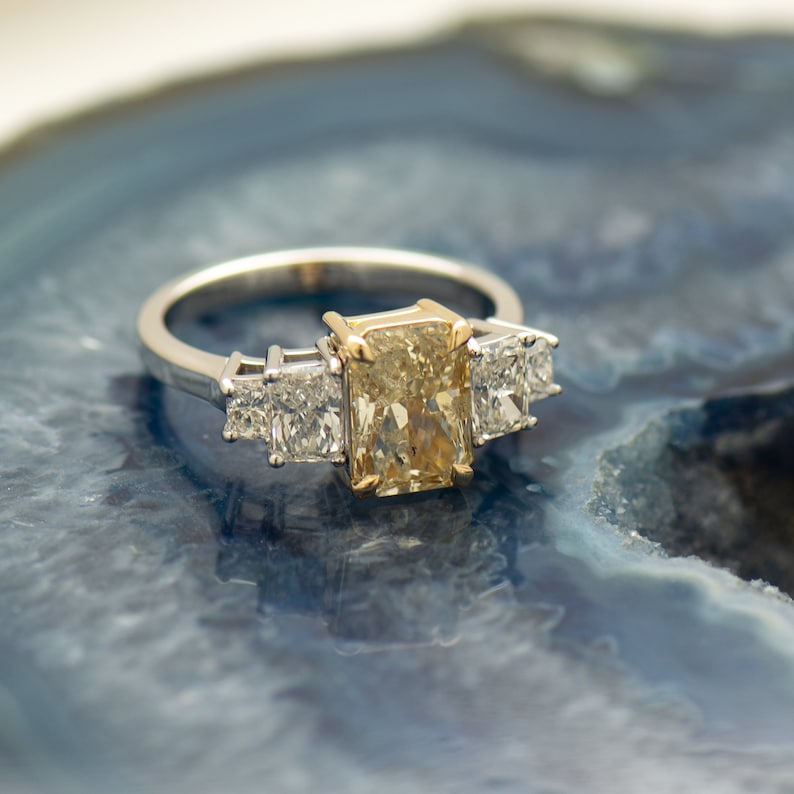 May include: A gold and white engagement ring with a large, rectangular, yellow diamond in the center and two smaller, square, white diamonds on either side.