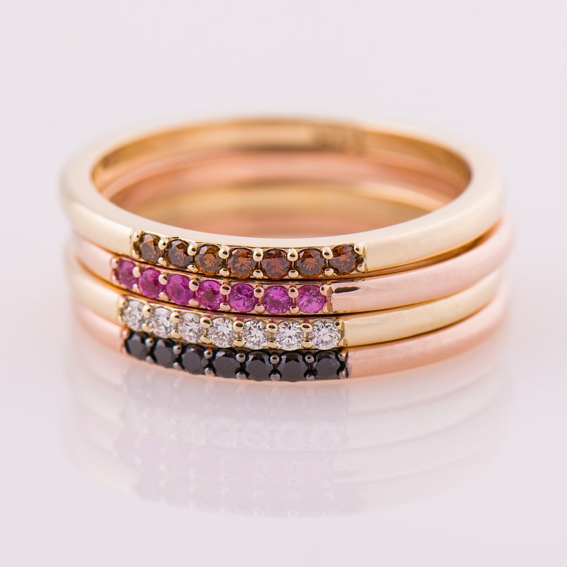 Peut inclure: Quatre bagues empilables en or avec des pierres pr&eacute;cieuses de diff&eacute;rentes couleurs. La bague du haut a des pierres brunes, la deuxi&egrave;me bague a des pierres roses, la troisi&egrave;me bague a des pierres claires et la bague du bas a des pierres noires.