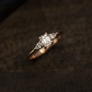 May include: A gold engagement ring with a large emerald cut diamond and two smaller pear-shaped diamonds on the band.