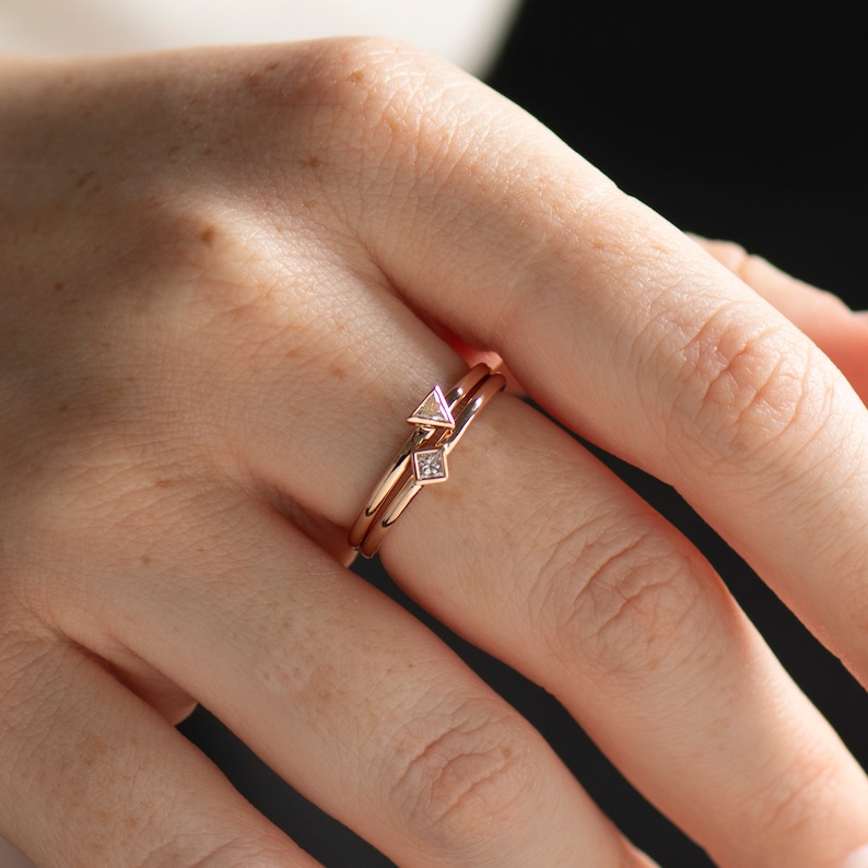 May include: Two delicate gold rings with small diamonds. The ring on top has a triangular diamond, and the ring on the bottom has a square diamond.