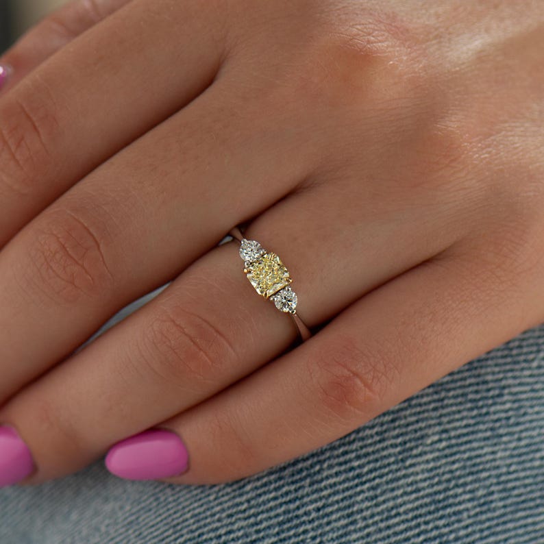 K&ouml;nnte beinhalten: Ein dreisteiniger Ring mit einem zentralen gelben Diamanten und zwei kleineren seitlichen Diamanten. Der Ring ist in Wei&szlig;gold gefasst.