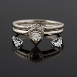 May include: A silver ring set with a triangular diamond, surrounded by smaller diamonds. The ring is on a black surface with two smaller diamonds on either side.