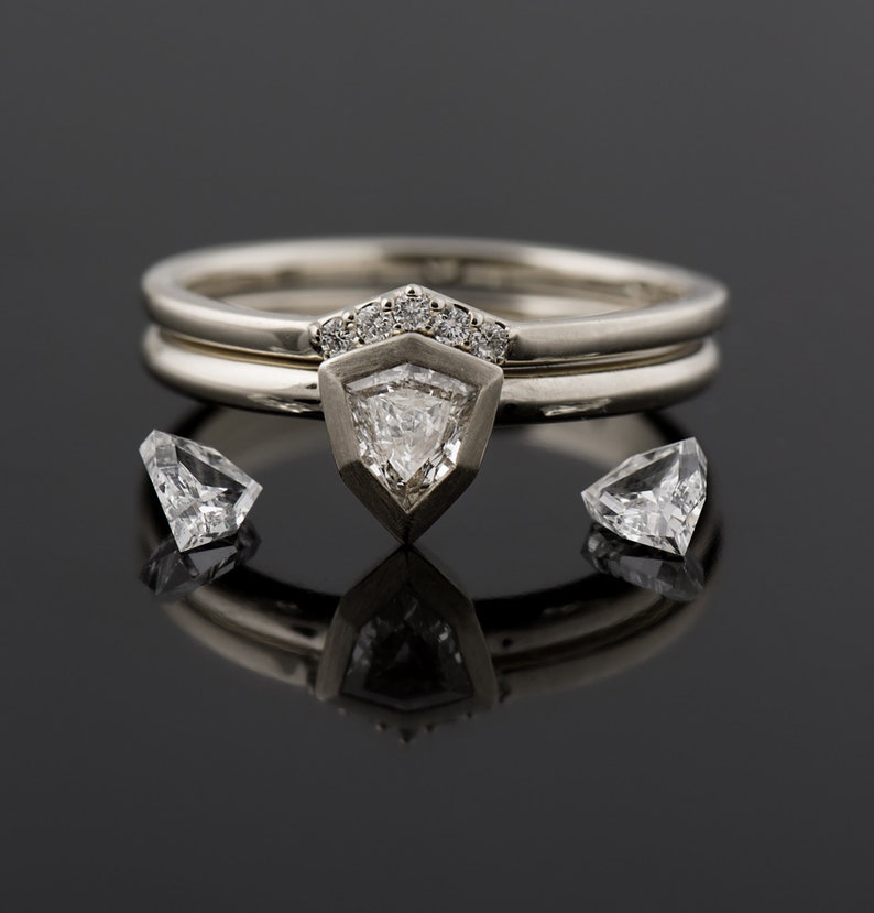 May include: A silver ring set with a triangular diamond, surrounded by smaller diamonds. The ring is on a black surface with two smaller diamonds on either side.