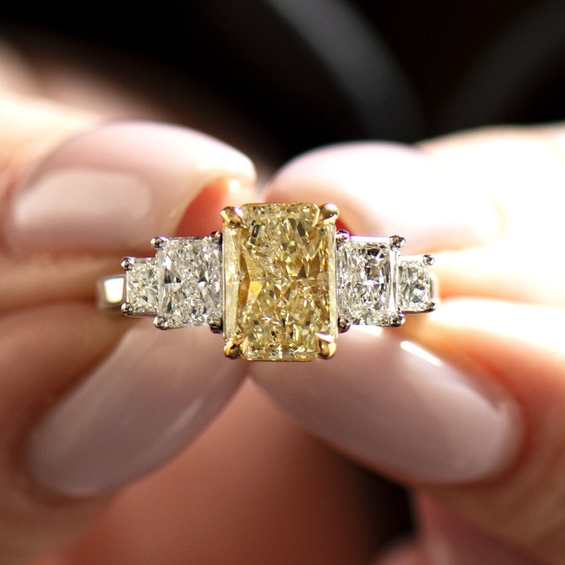 May include: A gold engagement ring with a large, rectangular, canary yellow diamond surrounded by two smaller, square-cut diamonds.