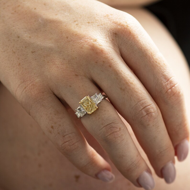 May include: A gold ring with a large, rectangular, yellow diamond in the center. The ring has two smaller, square, white diamonds on either side of the center stone.
