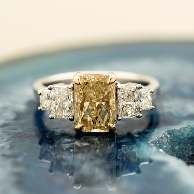 May include: A gold and white engagement ring with a large, rectangular, yellow diamond in the center and two smaller, square, white diamonds on either side.