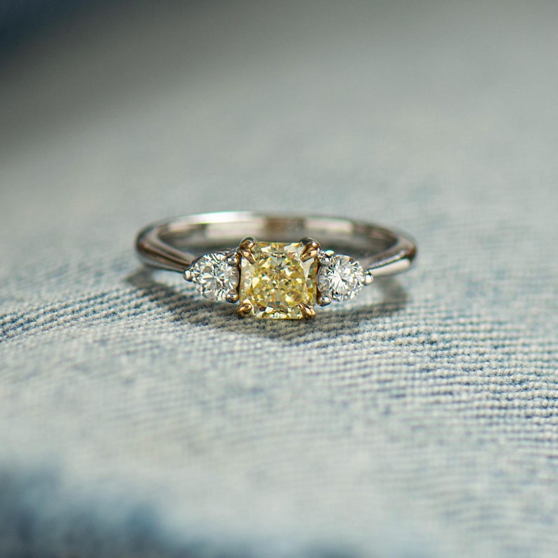 K&ouml;nnte beinhalten: Ein dreisteiniger Verlobungsring mit einem zentralen gelben Diamanten und zwei kleineren seitlichen Diamanten. Der Ring ist in Wei&szlig;gold gefasst.