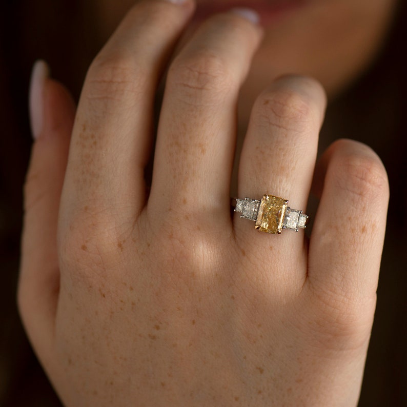 May include: A gold engagement ring with a large, rectangular, yellow diamond in the center. The ring is set with smaller diamonds on either side of the center stone.