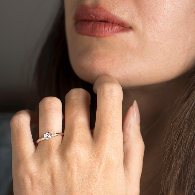Peut inclure: Une bague en or rose avec une pierre centrale ronde et transparente et de plus petites pierres d'accent. La bague est port&eacute;e au doigt, la main pr&egrave;s du menton. Un bijou d&eacute;licat.