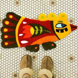 May include: A tufted rug shaped like a stylized bird in vibrant colors including yellow, red, orange, and dark green. The bird features a large blue eye, a floral pattern on its body, and a tail with five rounded sections, each containing a yellow circle.