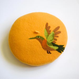 May include: Mustard yellow beret with an embroidered hummingbird design. The hummingbird features green, brown, and orange details. The beret is a round, soft hat, perfect for adding a touch of color and style to any outfit.