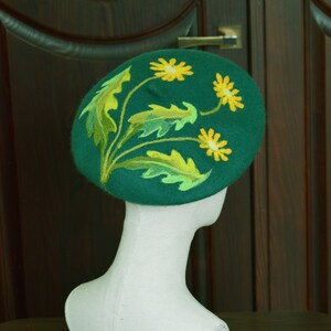 May include: A green felt beret with yellow embroidered flowers. The beret is on a white mannequin head.