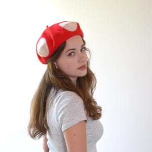 May include: A red beret with white circular accents, resembling a mushroom. The hat is made of a soft material and is worn by a person. The beret is a unique accessory, perfect for adding a touch of whimsy to any outfit.