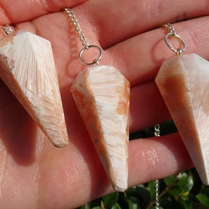 May include: Three orange and white crystal point pendulums with silver chains. The pendulums are hanging from the chains and are in the palm of a hand.