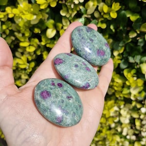 May include: Three oval gemstones are shown, each with a green base and red inclusions. The stones have a smooth, polished finish, giving them a natural look. The background is a blurred view of green leaves.