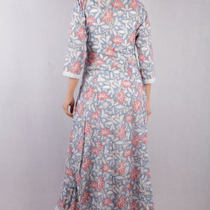 May include: A long, flowing grey dress with a floral print in shades of pink and white. The dress has a V-neckline and three-quarter sleeves. The model is wearing the dress with a pair of beige heels.