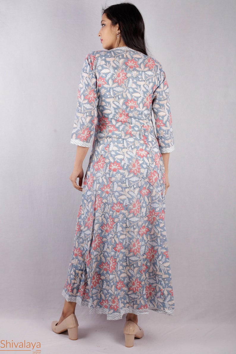 May include: A long, flowing grey dress with a floral print in shades of pink and white. The dress has a V-neckline and three-quarter sleeves. The model is wearing the dress with a pair of beige heels.
