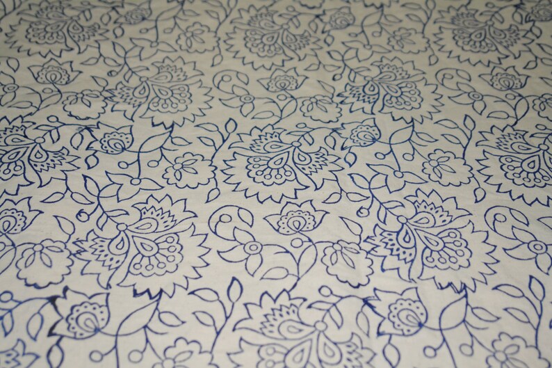May include: Close-up of a white fabric with a repeating pattern of blue floral designs. The pattern includes stylized flowers, leaves, and vines, creating a detailed and decorative textile.