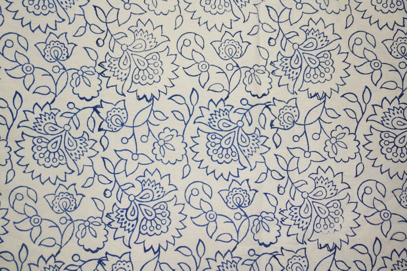 May include: Cream-colored fabric with a repeating pattern of blue floral designs. The pattern includes large, stylized flowers, smaller blossoms, and connecting vines with leaves. The design is evenly spaced across the fabric.