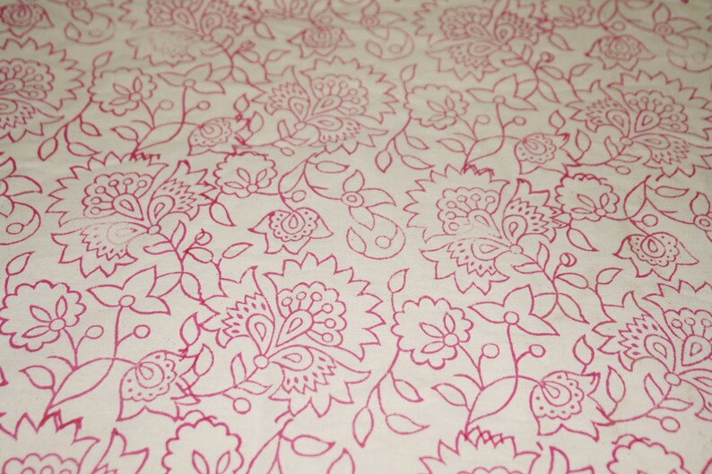 May include: Close-up of a cream-colored fabric with a repeating floral pattern in a vibrant pink hue. The design features stylized flowers, leaves, and vines, creating a delicate and intricate textile design.