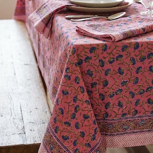 May include: A pink floral tablecloth with matching napkins, set on a wooden table. The tablecloth features a repeating pattern of blue flowers and a decorative border. Table setting includes plates, silverware, and glassware.