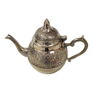 May include: A decorative, silver-colored teapot with intricate floral and leaf patterns etched across its surface. The teapot features a curved spout, a looped handle, and a conical lid with a pointed finial. The design is ornate and detailed.