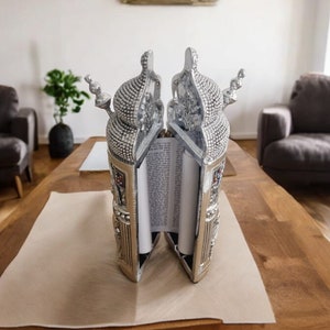 May include: A silver Torah scroll case with ornate details and a scroll of parchment inside. The case is open and the scroll is partially unrolled. The case is sitting on a wooden table with a white placemat.