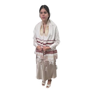 Op de afbeelding: Een witte tallit, een Joodse gebedssjaal, met bordeauxrode strepen en decoratieve gouden borduursels. De tallit wordt gedragen over een wit overhemd en een beige jurk. De sjaal heeft geknoopte franjes.