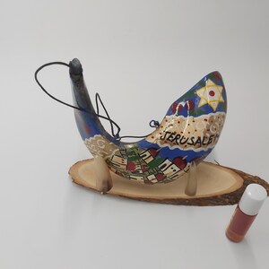 May include: A hand-painted shofar, a traditional Jewish ceremonial horn, with a blue and gold design depicting the city of Jerusalem. The shofar is on a wooden stand and a small bottle of oil is in the foreground.