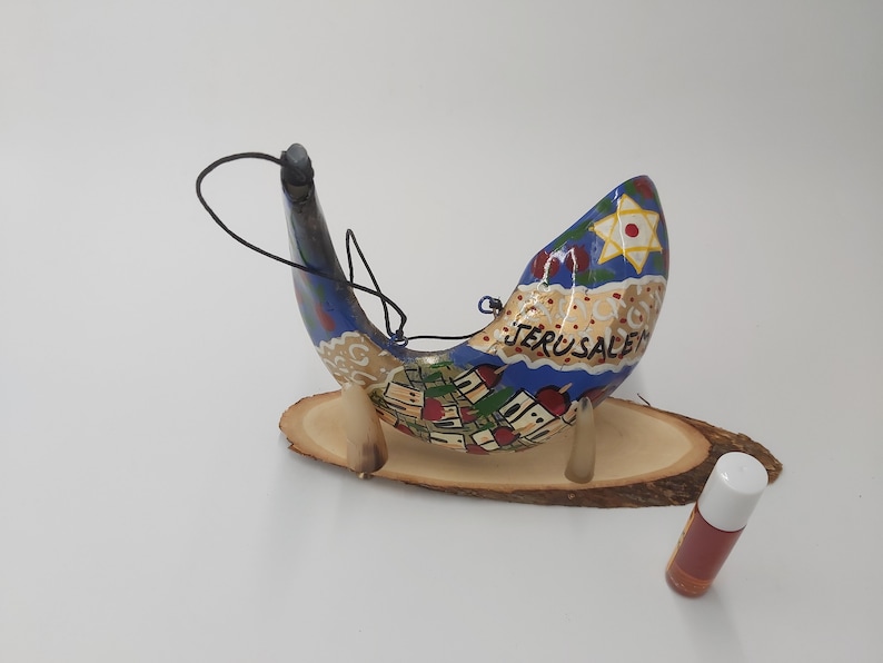 May include: A hand-painted shofar, a traditional Jewish ceremonial horn, with a blue and gold design depicting the city of Jerusalem. The shofar is on a wooden stand and a small bottle of oil is in the foreground.