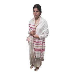 May include: A white tallit, a Jewish prayer shawl, with pink and gold stripes and fringe. The tallit is draped over a white shirt and beige pants. The garment has a traditional design and is worn by a person.
