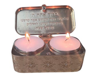 Pode incluir: Um castiçal de metal dourado com duas velas acesas. A tampa articulada está aberta, revelando texto gravado. A base apresenta um padrão floral decorativo. As velas são brancas e emitem um brilho quente.