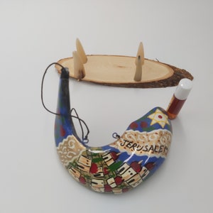 May include: A hand-painted shofar with a blue and gold design depicting the city of Jerusalem. The shofar is accompanied by a small bottle of oil and a wooden stand with three decorative horns.