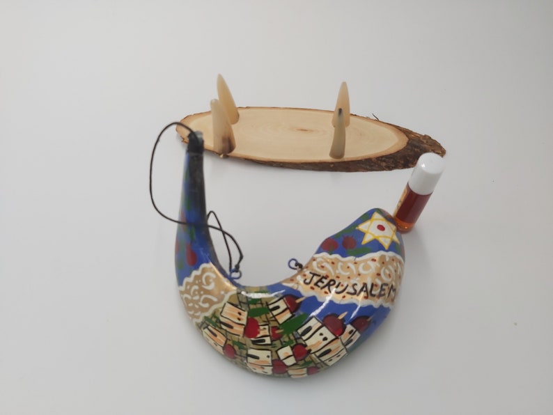 May include: A hand-painted shofar with a blue and gold design depicting the city of Jerusalem. The shofar is accompanied by a small bottle of oil and a wooden stand with three decorative horns.
