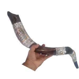May include: A brown shofar, a ram's horn, with silver and gold decorative bands. The bands feature intricate patterns and colorful gemstones. The shofar is held in a hand.