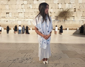 May include: A person wearing a white dress and a light blue and white scarf with a butterfly pattern. The person is standing in front of a large stone wall. Other people are visible in the background.