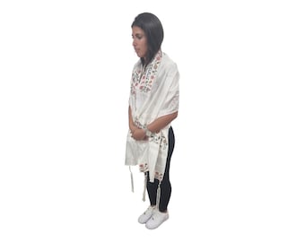 May include: A white prayer shawl with floral embroidery and tassels. The shawl is being worn by a person standing against a white background.