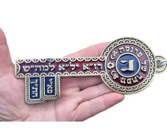 May include: A decorative key with Hebrew text, featuring a gold-colored metal frame and red and blue enamel details. The key has a circular head and a rectangular keyhole section, with a blue and white cord attached.
