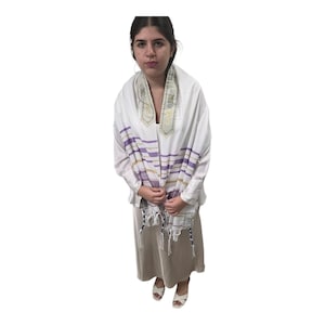 May include: A white prayer shawl with purple and gold stripes. The shawl is draped over the shoulders and chest. The garment has decorative embroidery at the neck and tassels at the bottom. The person is wearing a long beige dress and white sandals.