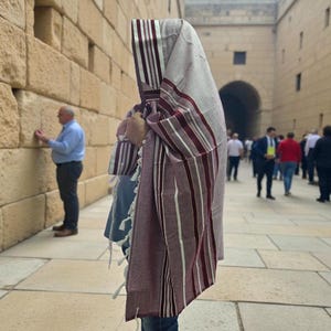 Op de afbeelding: Een persoon draagt een tallit, een Joodse gebedssjaal, met bordeauxrode en witte strepen. De sjaal is over het hoofd en de schouders gedrapeerd. De persoon staat voor een stenen muur, met andere mensen op de achtergrond.