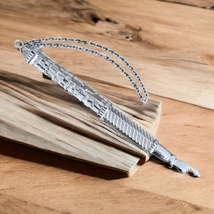 May include: A silver-toned yad, a Jewish religious pointer, with an intricate design and a chain attached. The yad is lying on a wooden surface.