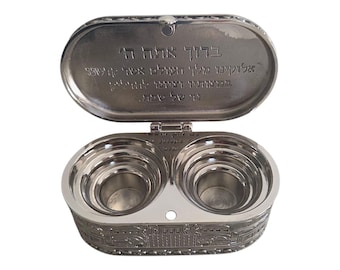 Pode incluir: Um recipiente prateado em forma oval com uma tampa aberta. O recipiente tem dois compartimentos aninhados e é gravado com texto hebraico na tampa. A base tem uma borda decorativa.