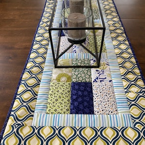 May include: A rectangular table runner with a patchwork design. The runner features a variety of fabrics in shades of blue, green, and white. A black metal candle holder with three glass cylinders and candles sits on top. The border has a navy blue and olive green pattern.