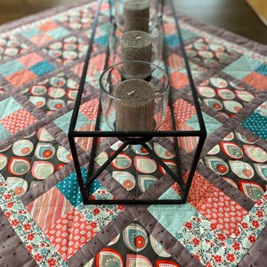 May include: A black metal candle holder with three gray candles on a patchwork quilt table runner with a variety of colors and patterns.