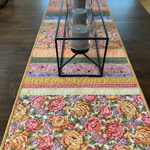 May include: A quilted floral table runner with a black metal candle holder. The runner features a repeating pattern of pink, peach, and yellow roses on a cream background. The candle holder holds three gray pillar candles in glass holders.